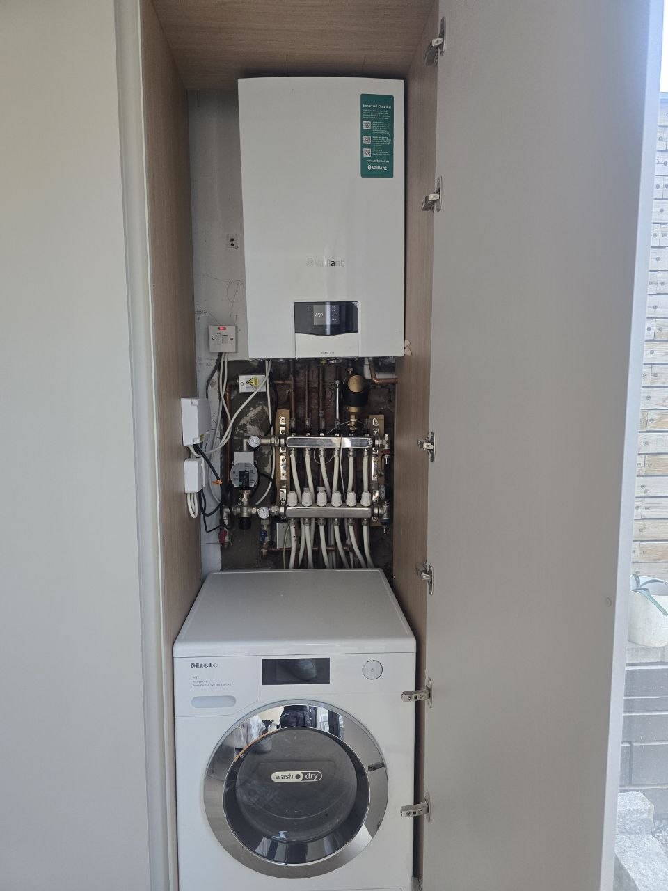 Vaillant boiler and underfloor heating manifold installed above a Miele washer dryer in a utility cupboard.