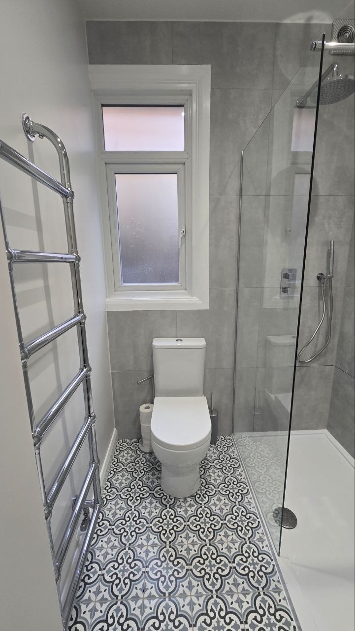 Walk-in shower beside a toilet with large grey wall tiles and patterned floor tiles.