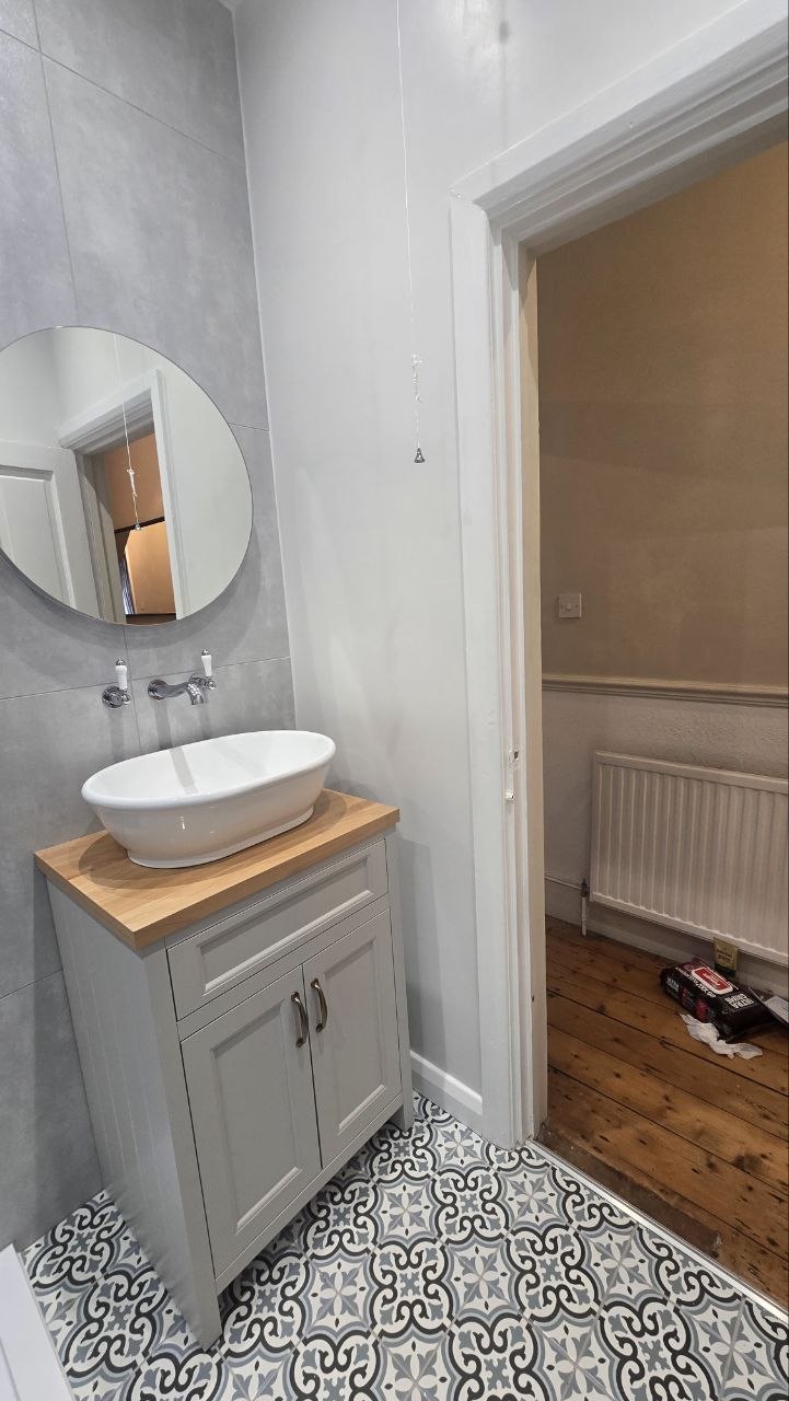 Bathroom vanity with white basin, timber-effect worktop, round mirror and patterned tiled floor.