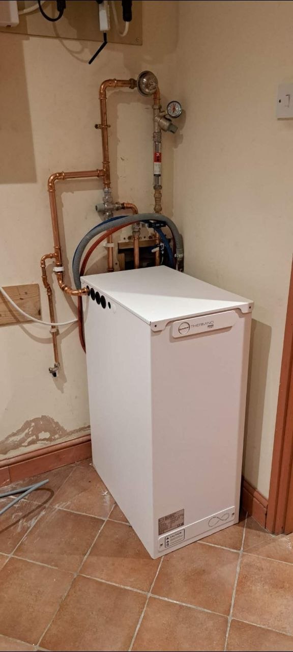 White heating boiler with copper pipework, valves and pressure gauge installed in a utility area.