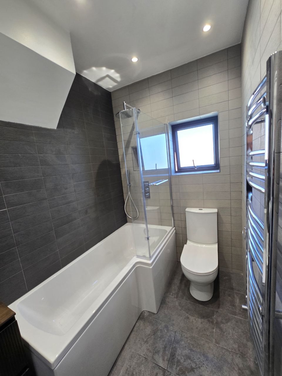 Modern bathroom with bath, glass shower screen, WC and grey wall and floor tiles in North London.