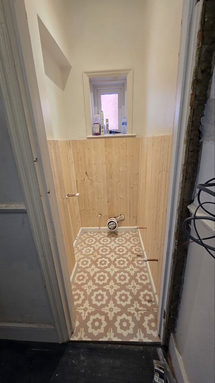 Cloakroom with finished patterned floor tiles, timber wall panelling and exposed plumbing ready for final installation.
