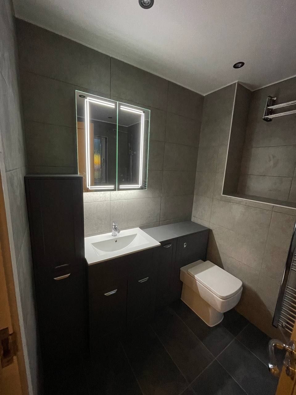 Modern renovated bathroom with grey wall tiles, dark vanity unit, WC and illuminated mirror.