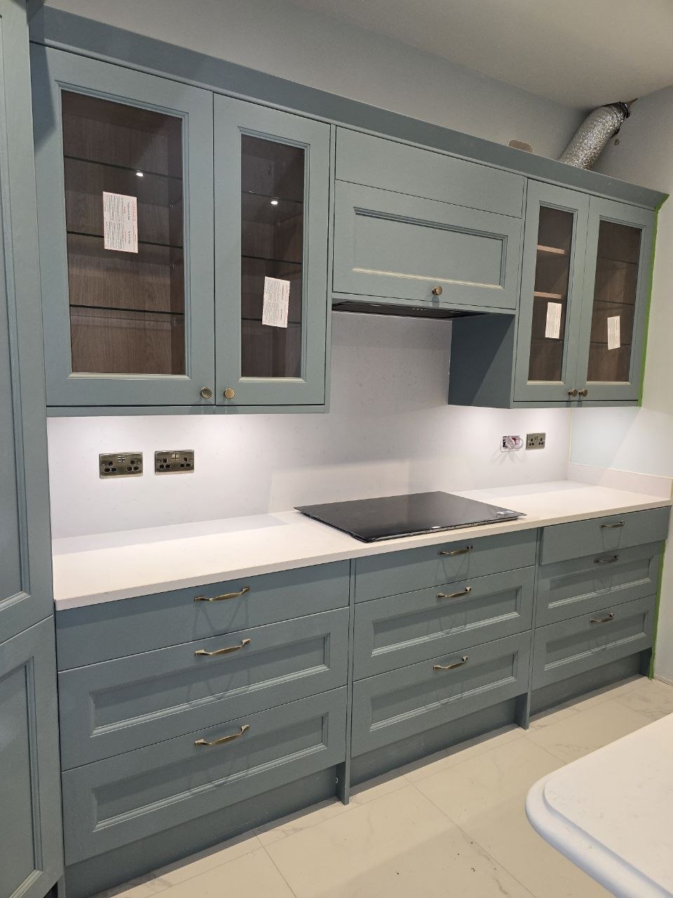 Blue-green shaker kitchen cabinets with white worktops, glass-fronted cupboards and a black induction hob.