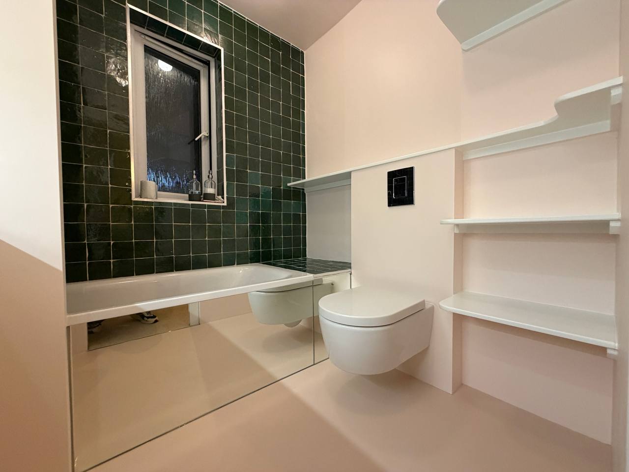 Modern bathroom with dark green wall tiles, fitted bathtub, wall-hung toilet, mirrored bath panels and white shelving.