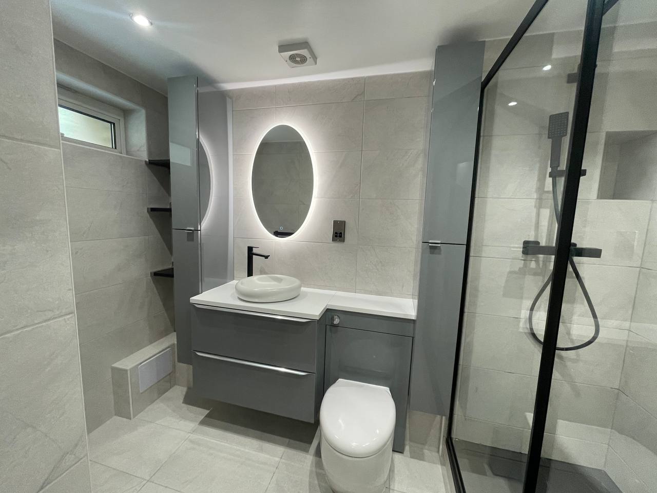 Modern bathroom showing grey vanity unit, illuminated oval mirror, WC and black-framed shower screen.
