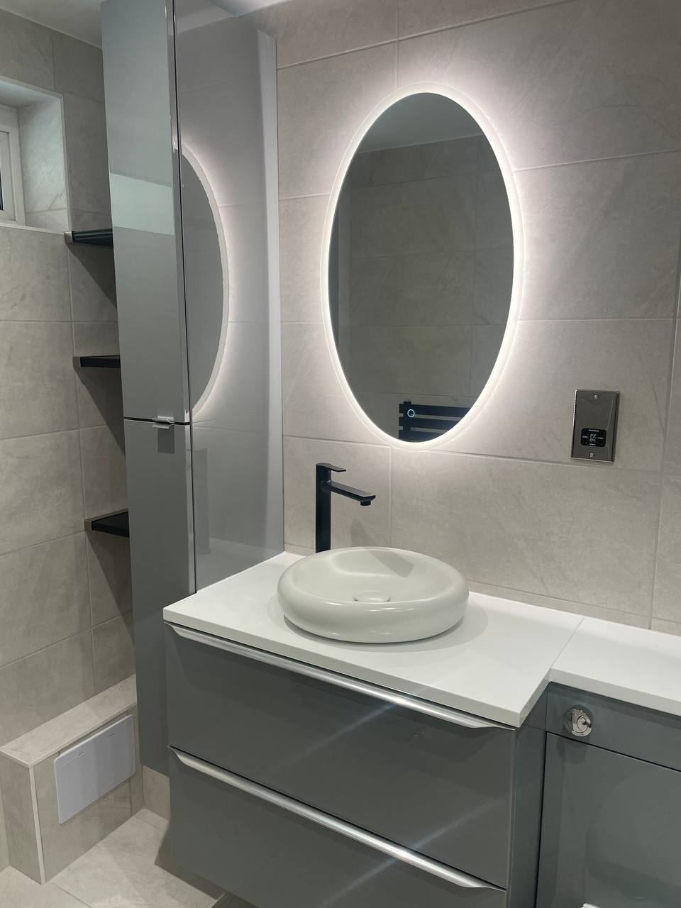 Close-up of grey bathroom vanity, white countertop basin, black tap and illuminated oval mirror.