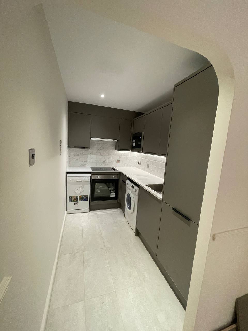 Modern fitted kitchen with dark grey cabinetry, white worktops and white tiled splashback.
