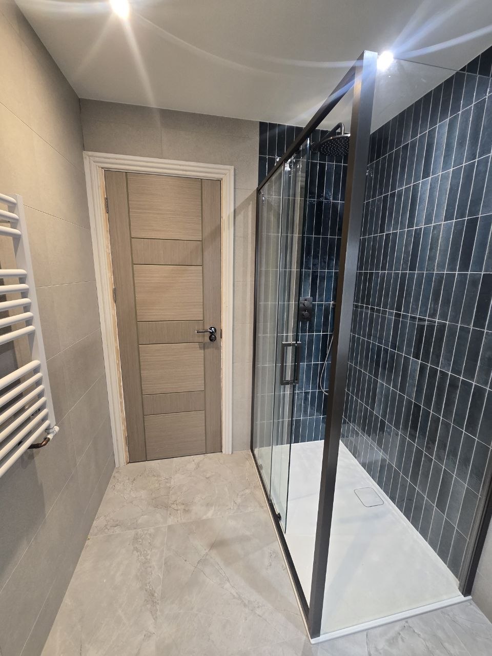 Modern bathroom with a walk-in shower, dark blue tiles, glass enclosure and marble-effect floor tiles.